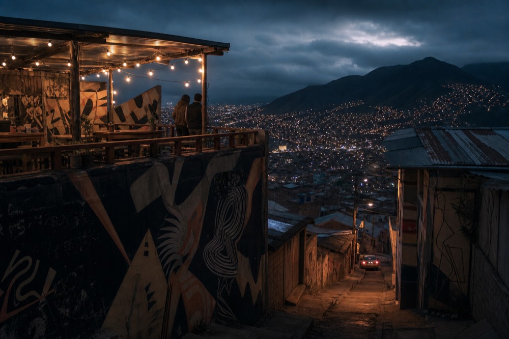 A rooftop terrace bar at dusk with string lights and low tables, two distant figures leaning on the rail above a valley of glowing windows