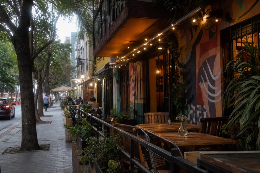 A leafy urban street of low-rise cafes with outdoor tables under trees, warm late-afternoon light, a few distant pedestrians with their backs to the camera