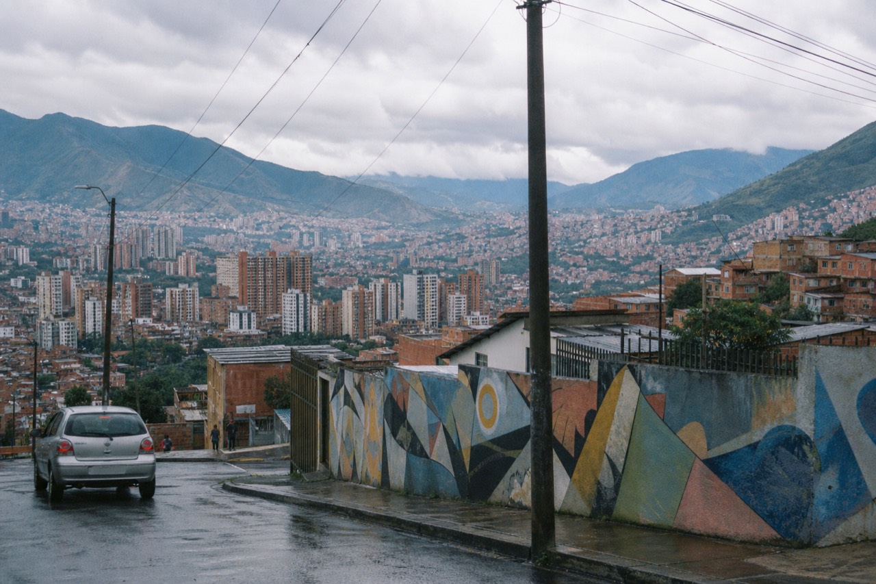 A wide daylight view over a green valley city of high-rise towers and red-brick hillsides, mountains rising on both sides under broken cloud