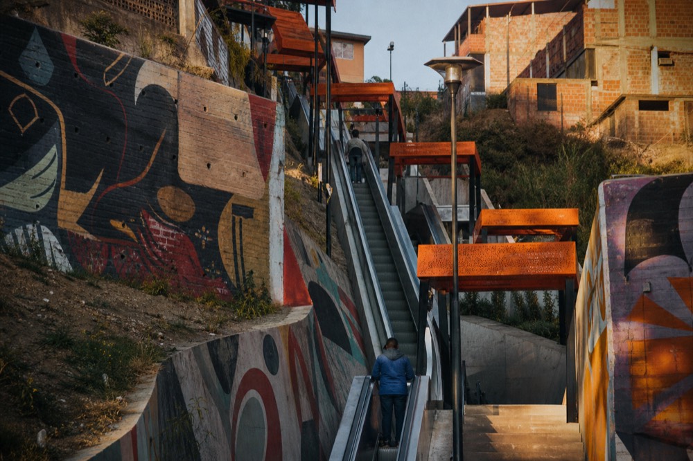 Outdoor public escalator sections climbing a steep hillside under orange canopies, painted retaining walls either side, soft morning light