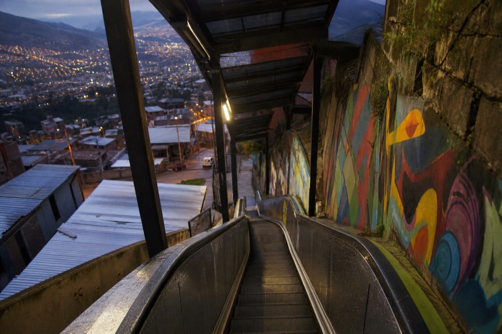 Rain-wet escalator steps under a canopy with water beading on the metal and grey light on the hillside houses beyond