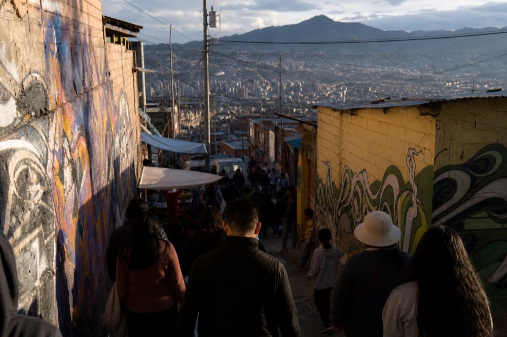 Crowd of visitors packed onto a hillside landing at midday seen from above and behind, painted walls and stalls on both sides