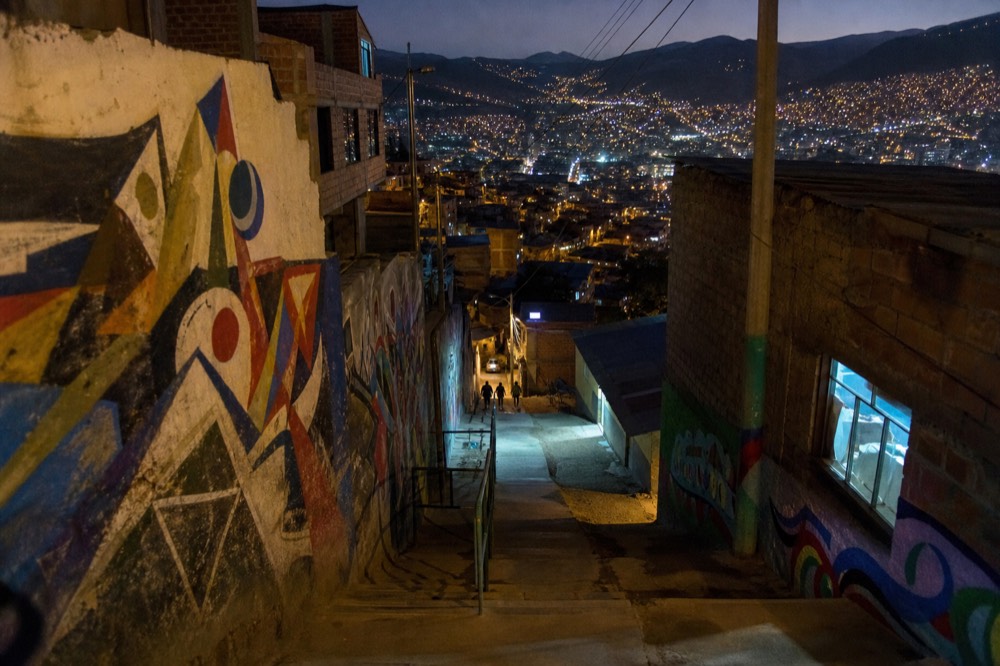 Stacked hillside houses glowing with window lights at dusk seen from high on the slope with the dark valley spread below