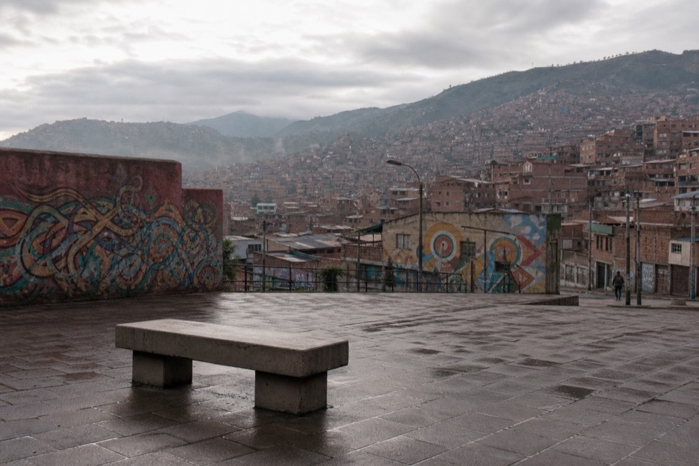 Empty hillside plaza at dawn with wet paving stones, a single concrete bench and rows of brick houses rising behind under grey cloud