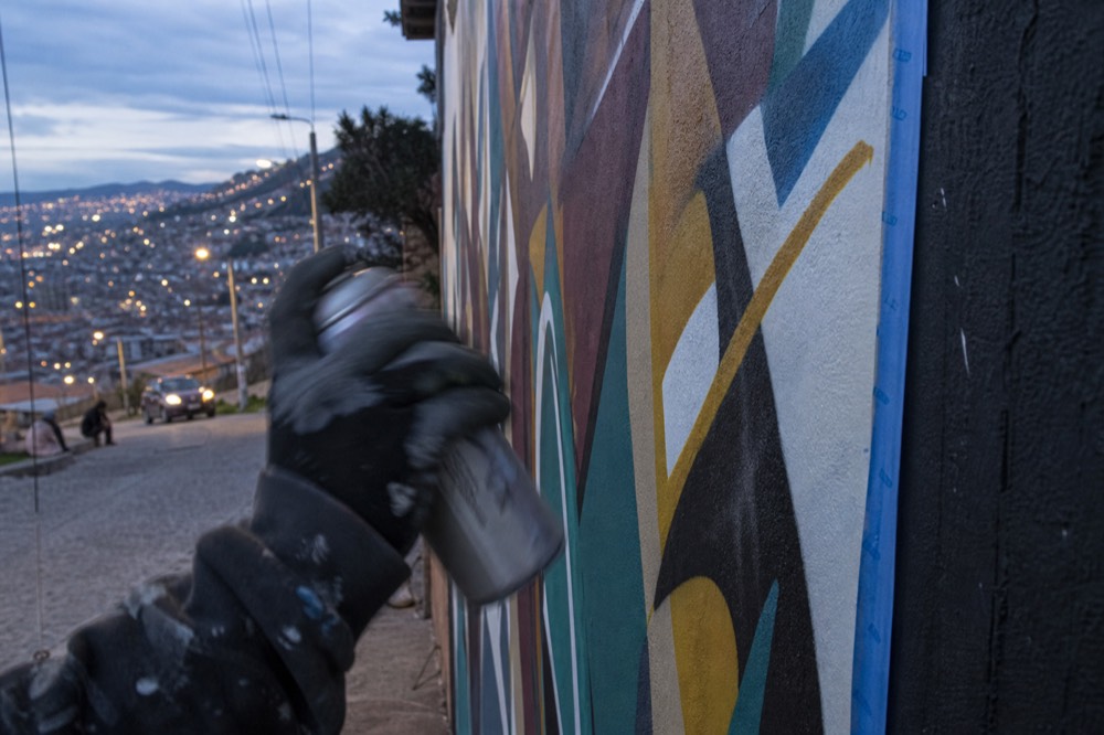 Painter's gloved hand shaking an unlabelled spray can in front of a half-finished abstract wall panel with masking tape along its edge