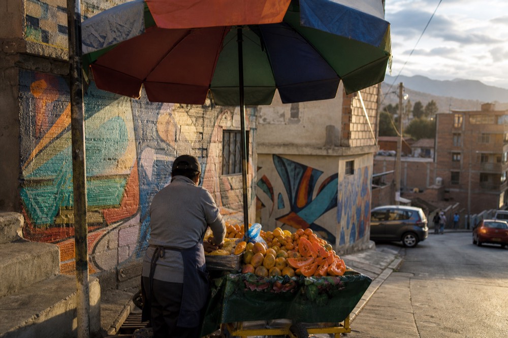 Fruit cart piled with mango, papaya and lulo on a sloping street corner, vendor slicing under a bright umbrella