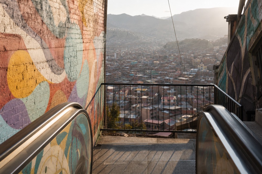 Empty landing between two outdoor escalator flights with a metal railing overlooking a valley of dense rooftops, abstract painted mural filling the side wall