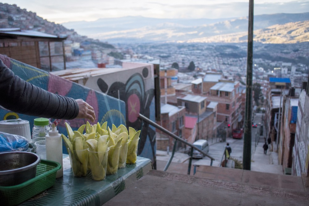 Street stall on a stair landing with cut green mango in plastic cups, a hand reaching for one, painted wall and hillside houses behind