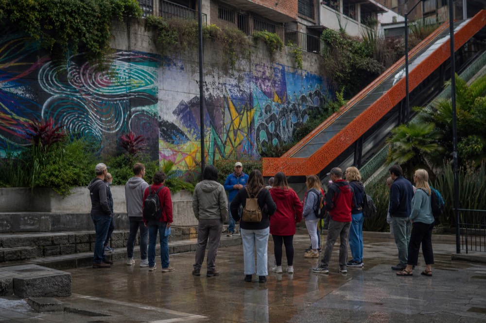 A dozen visitors standing loosely around a guide at a small paved plaza beside an orange-roofed outdoor escalator, colourful abstract wall art behind them