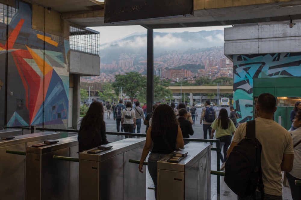 A ticket barrier and turnstiles at a busy Medellin metro station entrance, commuters passing through, daylight flooding in from the street beyond