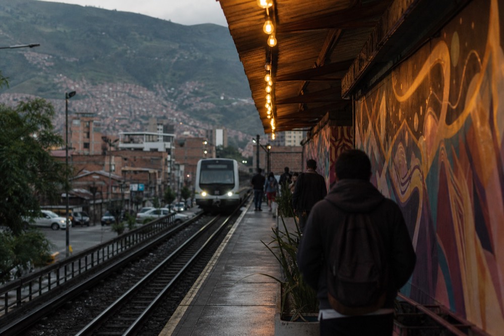 Silver commuter train pulling into an elevated open-air platform, a few distant passengers waiting, green hillside neighbourhoods visible beyond the tracks