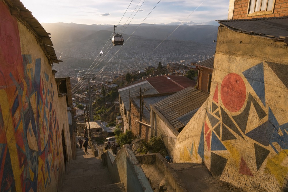 Small enclosed cable-car cabin gliding on a steel line above corrugated rooftops on a steep green slope, hazy valley behind