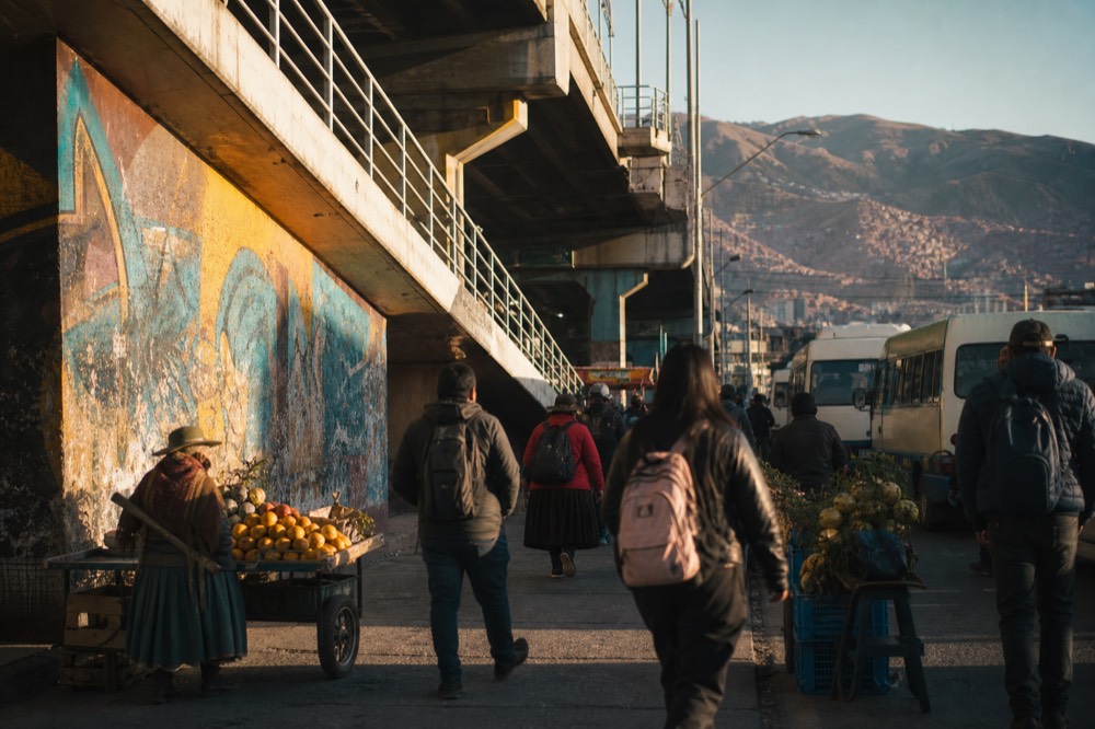 Busy plaza outside an elevated station with fruit carts, waiting minibuses and travellers seen from behind in warm morning light