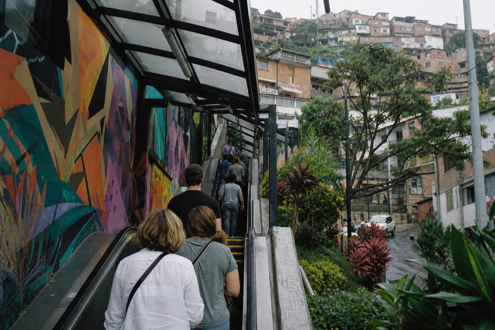 A small group of visitors photographed from behind following a guide up a set of covered outdoor escalators, hillside houses rising steeply on both sides